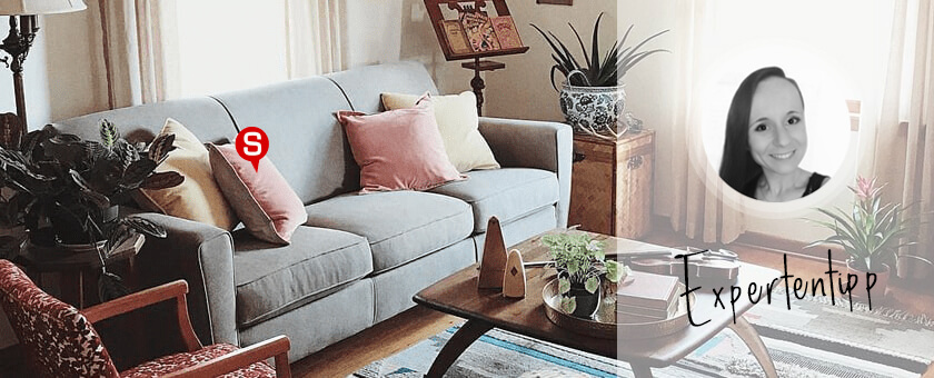 Ein gemütliches Wohnzimmer im Vintage-Stil mit einem grauen Sofa, einem hölzernen Couchtisch und rosa Elementen. Auf dem Sofa liegen Dekokissen in Pastellfarben. Das Gesamtbild wird von einem folkloristischen Teppich auf dem Holzboden abgerundet.