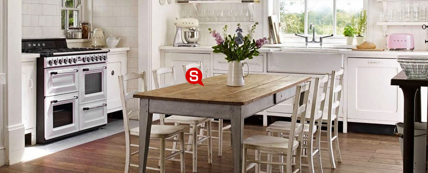 Ein gemütliches, helles Esszimmer im Vintage Stil mit einem hölzernen, alt gemachten Tisch in der Mitte des Raumes. Auf dem Tisch steht eine weiße Vase mit bunten Blumen und um den Tisch stehen sechs Holzstühle.