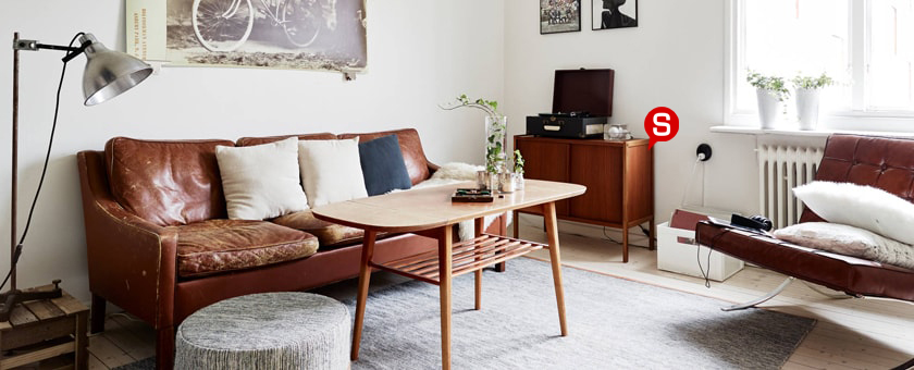 Ein stimmungsvolles, in Weißtönen gehaltenes Wohnzimmer mit Sitzmöbeln aus Leder. Auf den braunen Ledersofas liegen helle Dekokissen. Auf dem Couchtisch steht eine Glasvase mit Pflanzen und unter dem Tisch liegt ein moderner, grauer Teppich.
