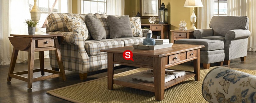Ein Wohnzimmer im Landhausstil mit kariertem Sofa und Holzmöbeln. Auf dem Sofa liegen Dekokissen und daneben steht ein Couchtisch. Das Gesamtbild wird von safrangelben Wänden und einem großen Teppich abgerundet.