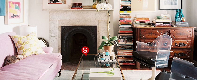 Eine Einrichtung im Vintage-Stil mit einem rosa Sofa und einem Couchtisch aus Holz. Auf dem Sofa liegt ein Dekokissen und auf dem Couchtisch steht eine Pflanze im Topf. Im Hintergrund ist eine Kommode aus Holz und zahlreiche Bücher neben dem Kamin zu sehen.